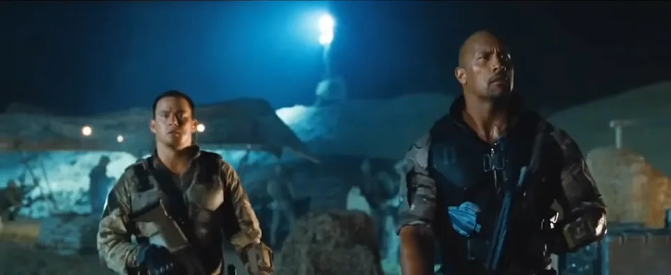 Two actors in tactical gear walk through a dimly lit outdoor setting, appearing focused and ready for action