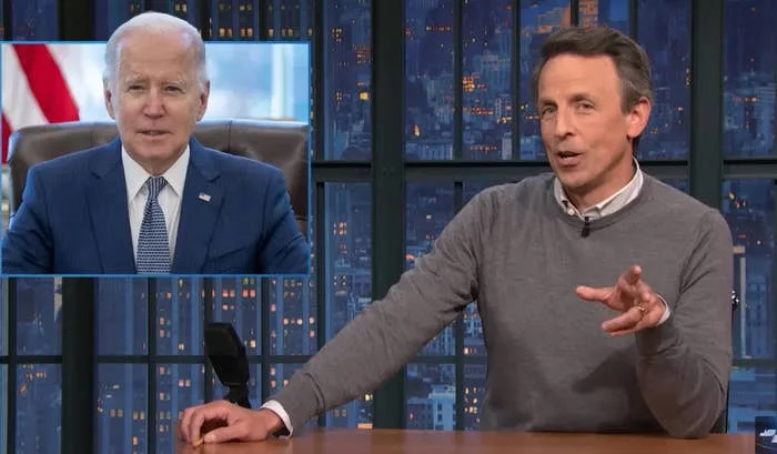 A talk show host sits at a desk, gesturing, with an image of a male world leader on a screen in the background