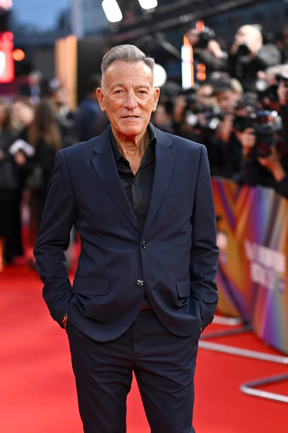 Person in a tailored suit on the red carpet, hands in pockets, with photographers in the background
