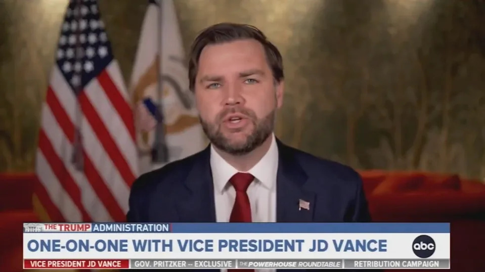 Man in suit and tie speaking in a news interview, with flags in the background. Lower third text labels him as Vice President JD Vance
