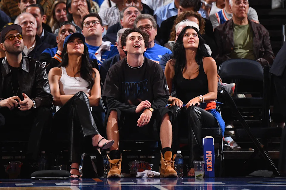 Three people sit courtside at a basketball game. The two women wear stylish outfits with leather elements; the man wears boots and casual clothing