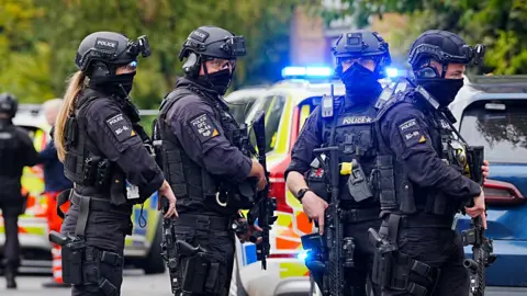 PA Media Armed police officers at the scene near the Heaton Park Hebrew Congregation synagogue in Crumpsall, Manchester, where two people died in a suspected terror attack by a man who was shot dead by police. The four officers are dressed in black, holding assault rifles and wearing helmets with face coverings drawn up over their mouths. 