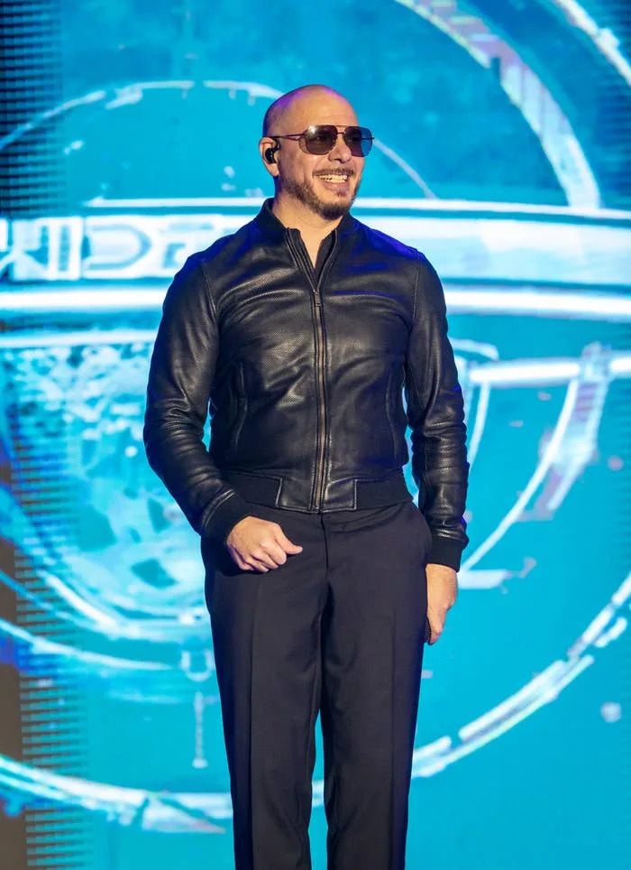 A man in sunglasses and a sleek leather jacket smiles on stage, set against a brightly lit backdrop with geometric designs
