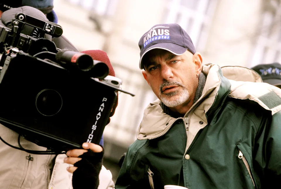 Person wearing a cap and a thick jacket stands behind a camera outdoors, focused and possibly involved in a film production