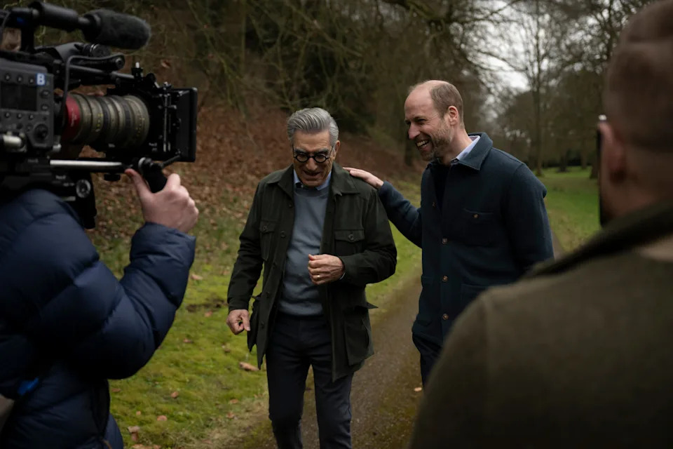 Prince William putting his hand on Eugene Levy's shoulder walking in the park while a cameraman films him