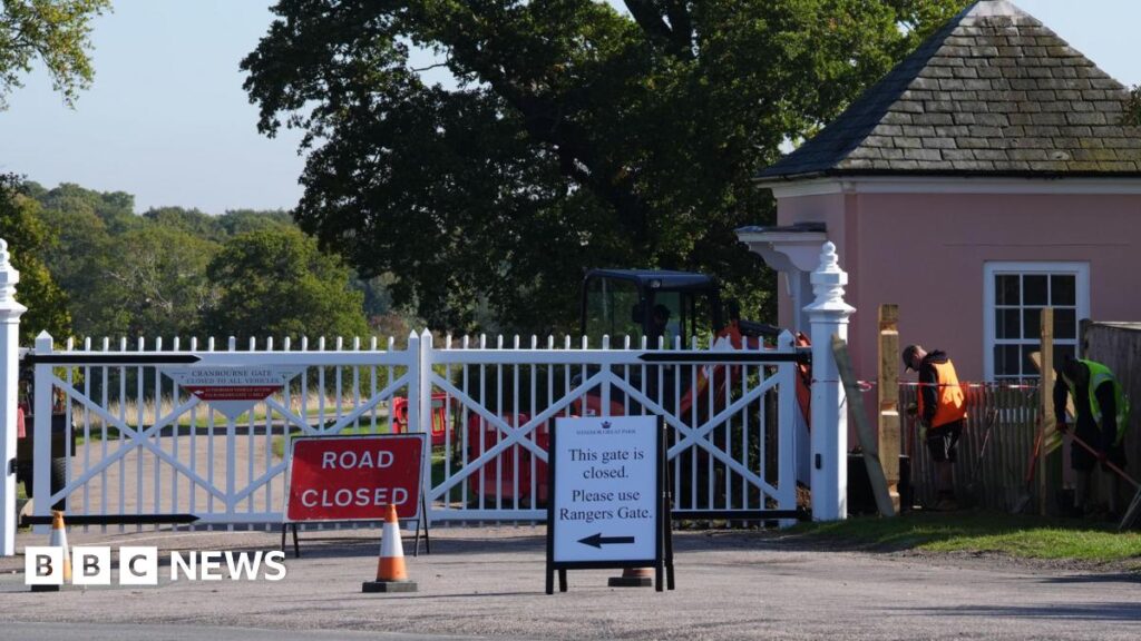 A white gate, Cranbourne Gate, in Windsor Great Park, with two workmen stood to the right of it. One is wearing an orange jacket and the other a green jacket. There is a red "road closed" sign in front of the gate and a sign to its right which says: "This gate is closed, please use Rangers Gate".