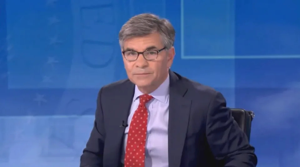 A seated man in a suit and red tie, appearing to host a news program in a studio setting