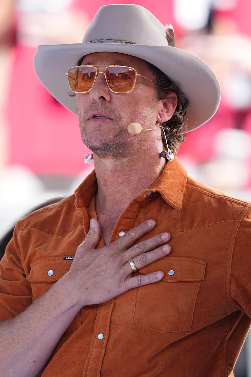 Person wearing a hat and sunglasses with a hand on their chest, speaking into a microphone