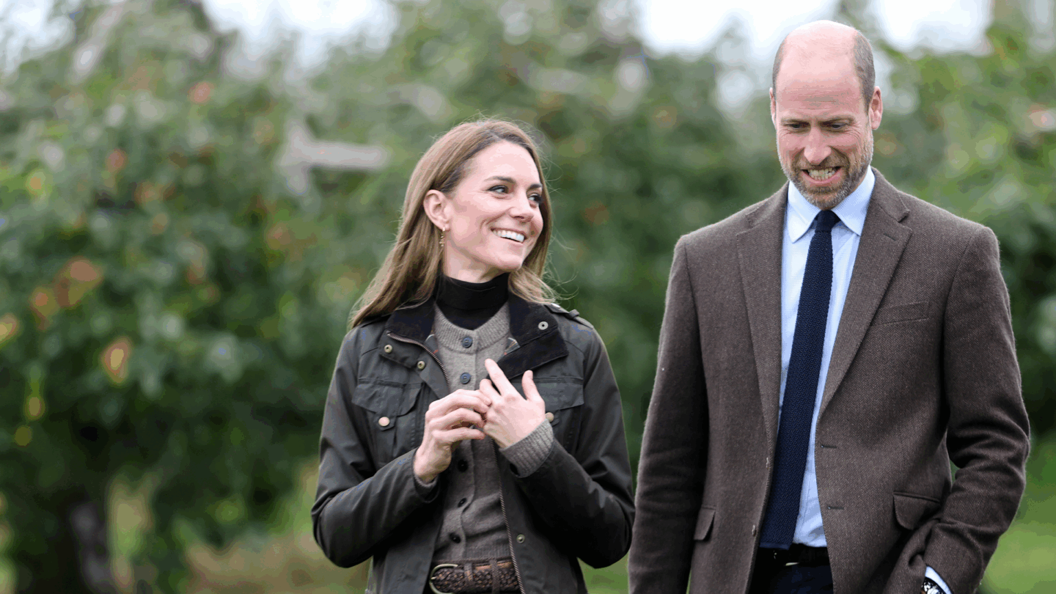 Kate Middleton wearing a green coat and brown cardigan walking next to Prince William in an orchard