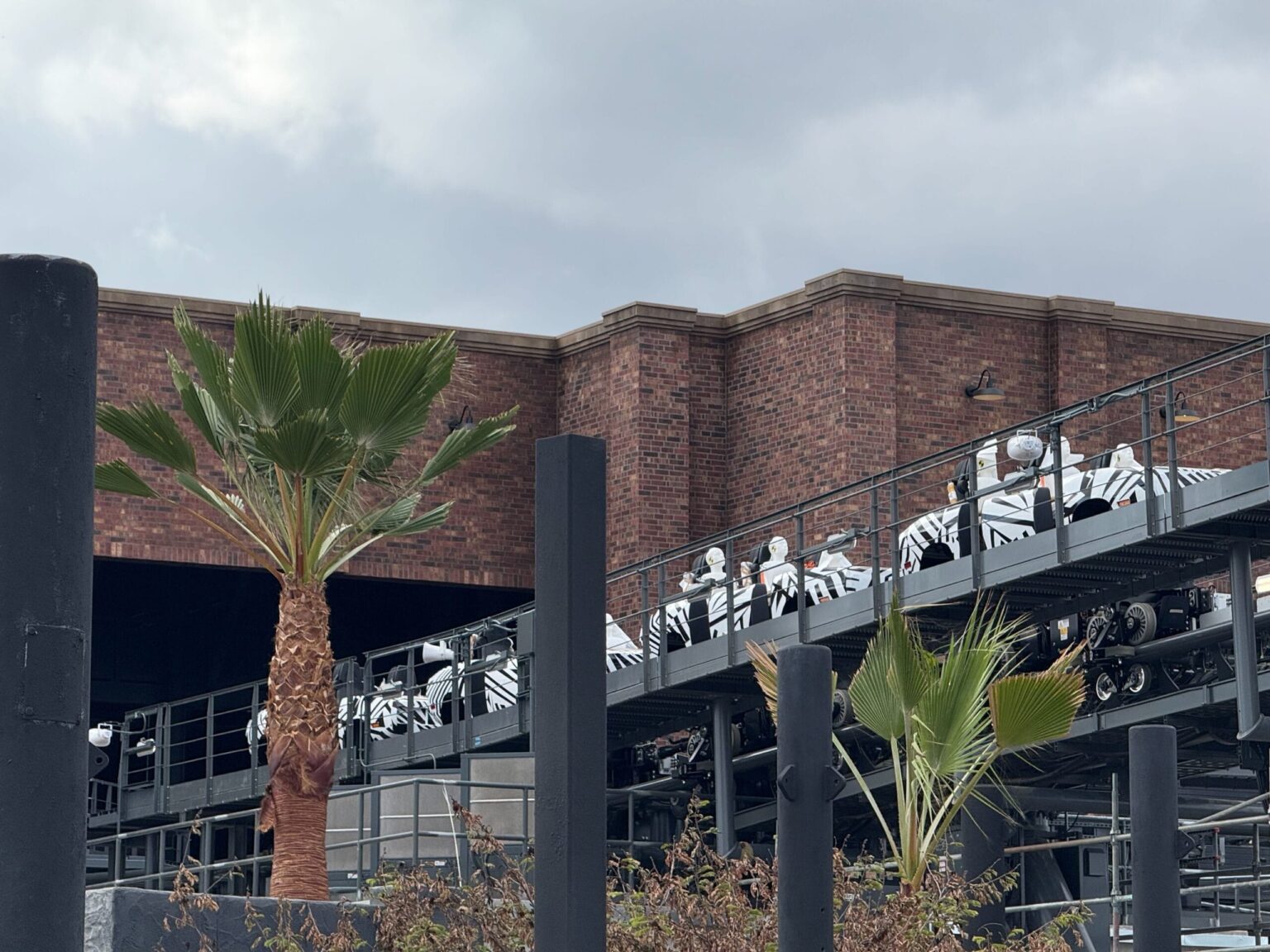People in white-and-black uniforms ride a roller coaster near a brick building and palm trees, similar to Disney parks’ theming.