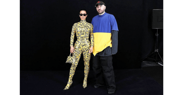 Kim Kardashian poses with Balenciaga’s former creative director Demna Gvasalia, wearing a yellow bodysuit covered in Balenciaga logo tape, standing against a black backdrop.