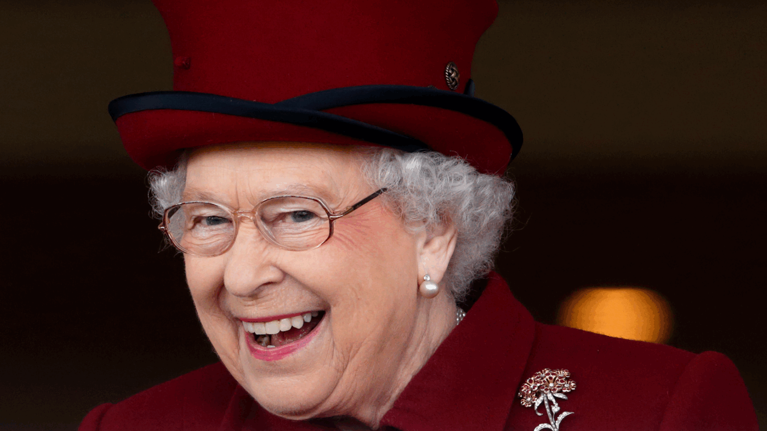 Queen Elizabeth Would Eat Four Meals a Day—And Would Ration One Thing “Until It Was All Gone” A headshot of Queen Elizabeth wearing a burgundy hat and coat while laughing