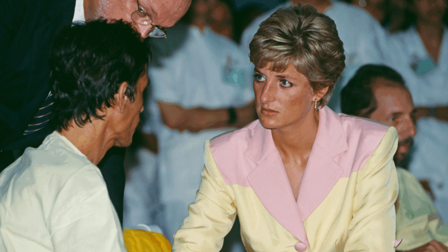 Princess Diana in a yellow and pink blazer talking to a man in a hospital gown