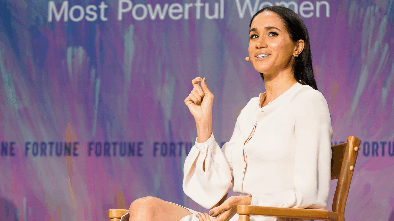 Meghan Markle Talks Business in an Ivory Blouse and Leather Pencil Skirt Meghan Markle wearing a white blouse sitting in front of a purple background
