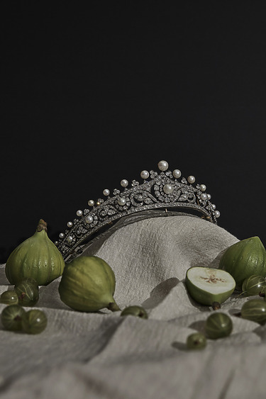 Silver tiara with pearls surrounded by green figs and gooseberries on beige fabric against a dark background.