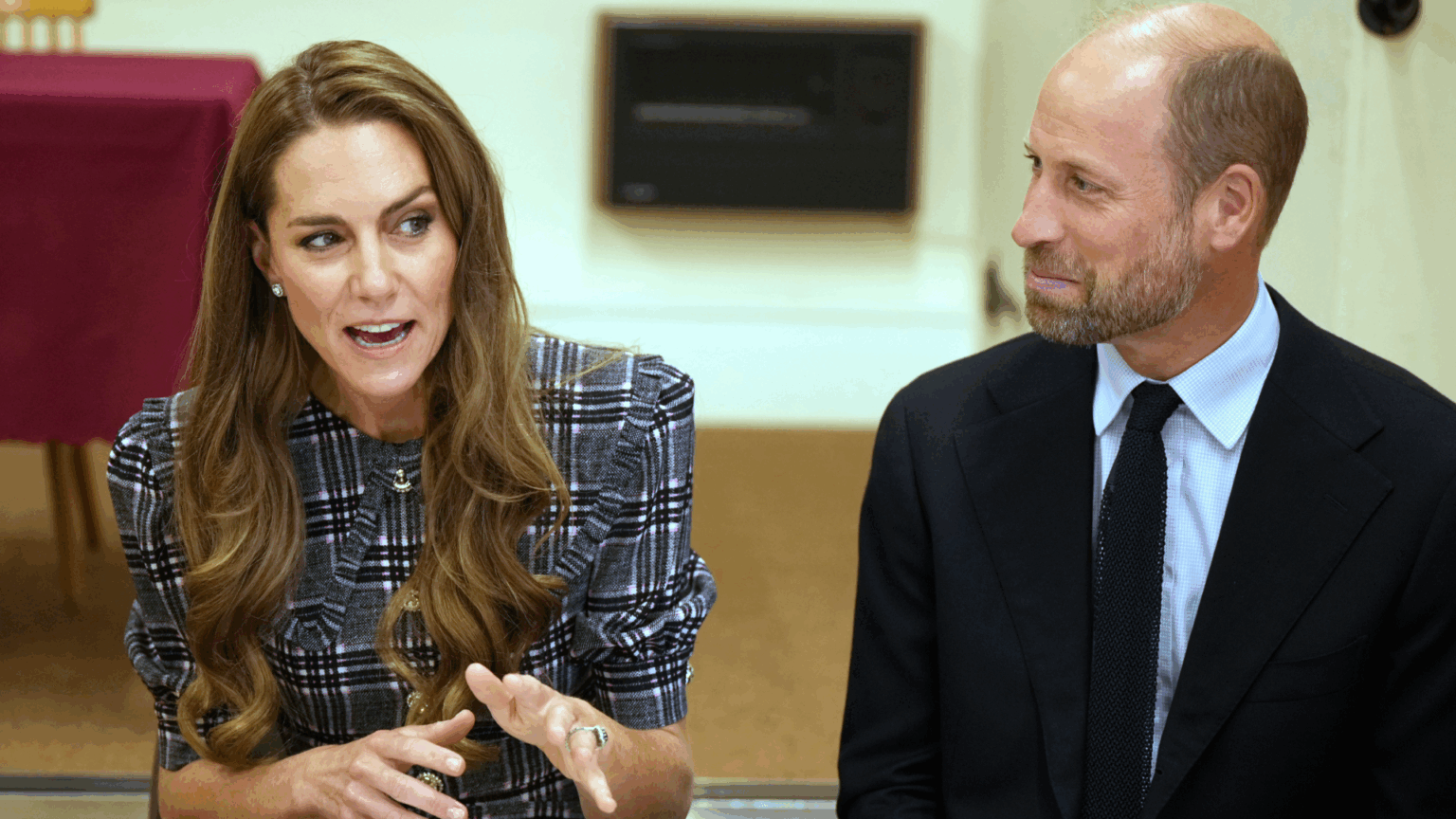 Prince William wearing a dark suit sitting next to Kate Middleton and looking at her, smiling, as she talks and gestures with her hands