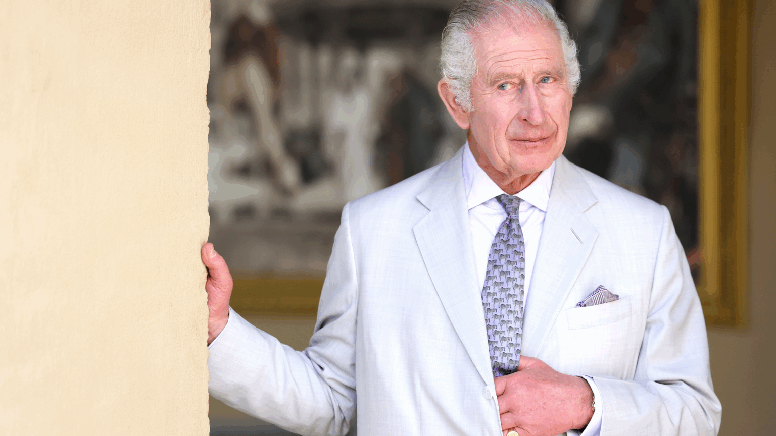King Charles Enters a New Era by Making a Huge Royal Change King Charles wears a light suit, and places his hand inside the jacket while leaning on a wall and looking mischievous