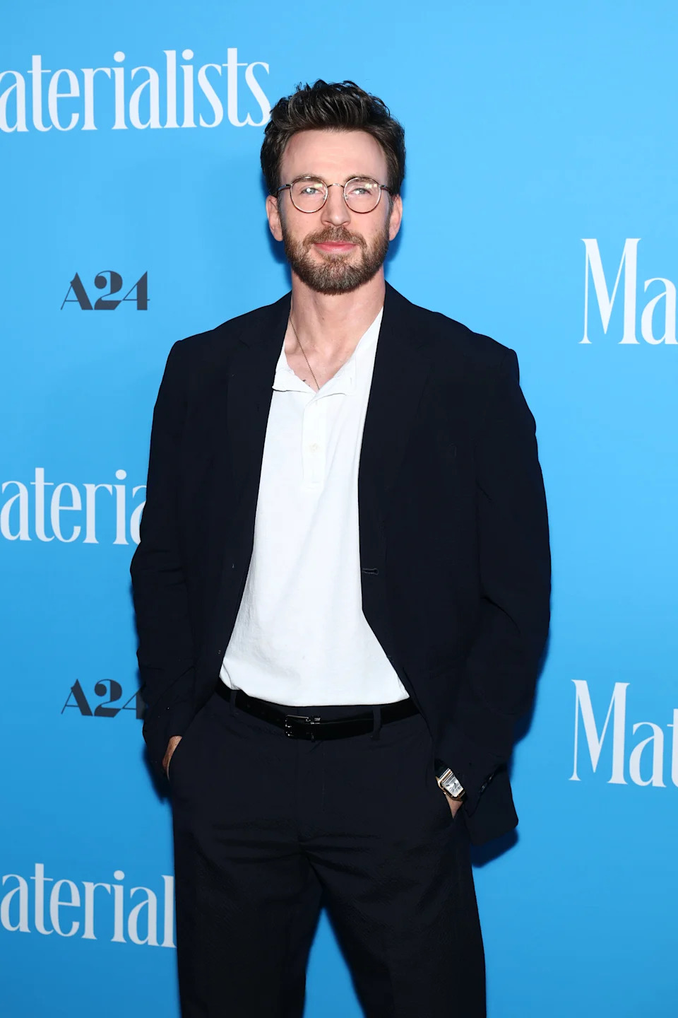 A man in a black suit with a white shirt and glasses poses on the red carpet at a "Materialists" event