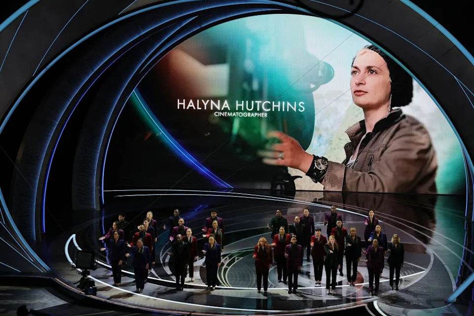 People on stage at a tribute for Halyna Hutchins, with her image displayed on a large screen in the background