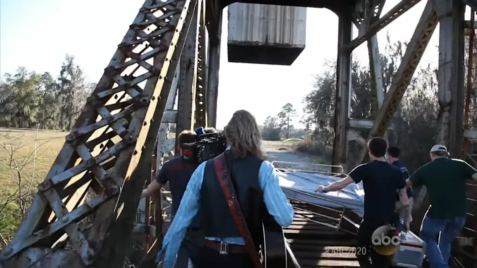 People walk across a metal bridge, some carrying gear, with a guitarist in front. Trees and fields are visible in the background