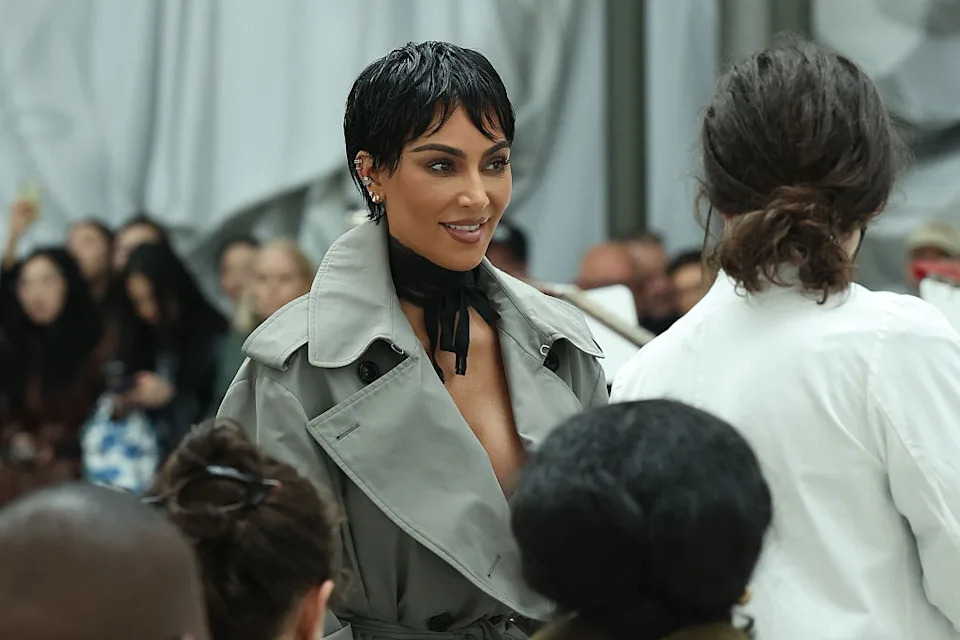 Person in a stylish trench coat with a black scarf, smiling at another person in a crowded indoor setting