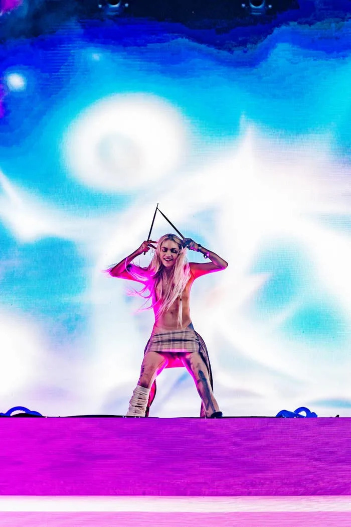 Performer on stage with long hair and plaid skirt, posing with arms raised, in front of an abstract light display