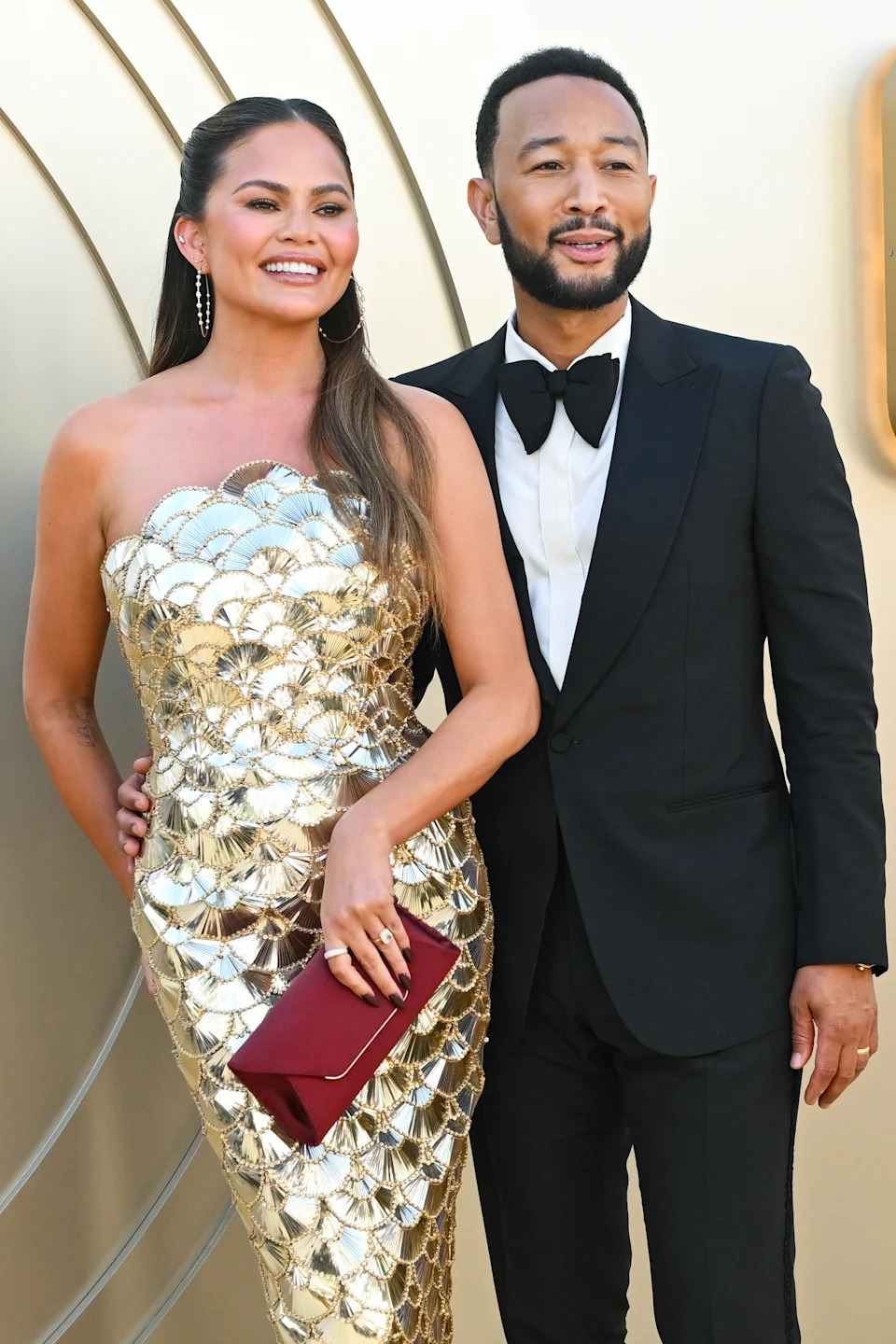 A woman in a strapless, metallic, patterned gown and a man in a tuxedo smile on the red carpet