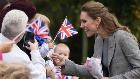 The Times/PA The Princess of Wales, wearing a grey suit and holding a small bunch of flowers, smiles as she meets a croud of people who are waving flags and taking photographs.