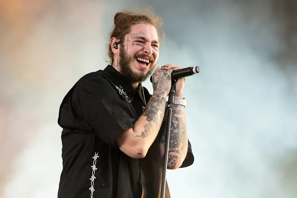 Smiling musician performing on stage, wearing a casual shirt with tattoos visible on arms. Holding a microphone, enjoying the performance