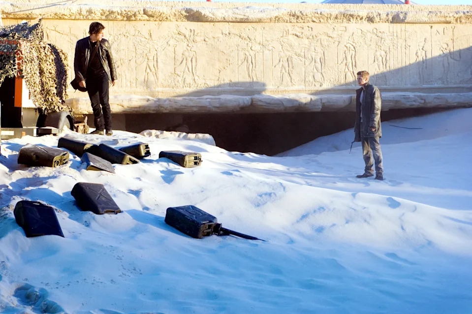 Two people stand on a snowy surface near a stone wall with carvings and scattered objects. They appear to be engaged in a tense situation