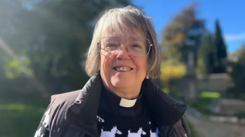 BBC Reverend Sue Murray from the Benefice of Bisley smiles at the camera in the churchyard with the sun shining. She wears a black jumper with white dogs on it with a gilet and a clerical collar. 