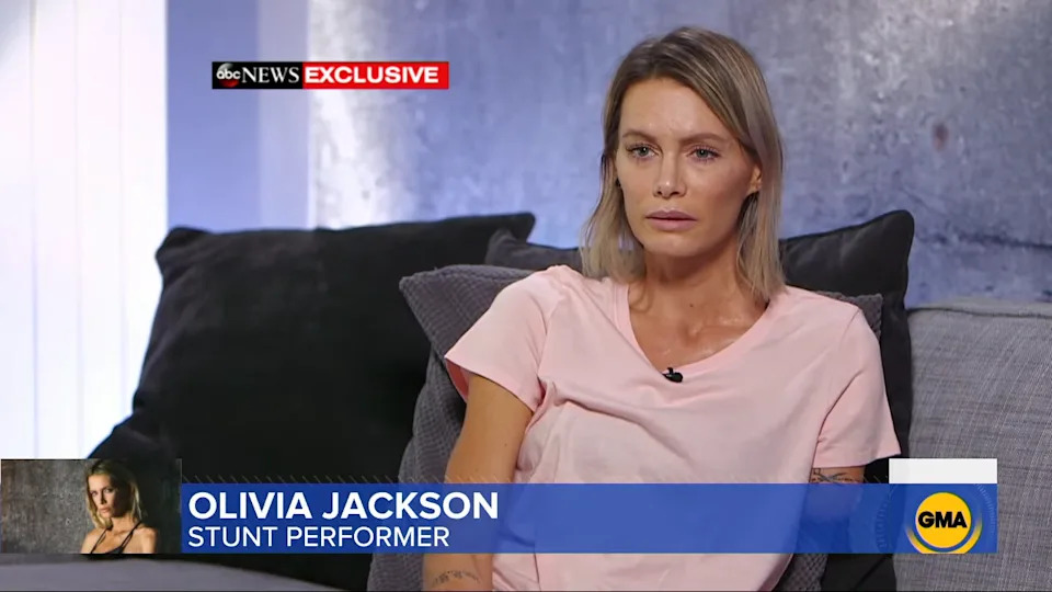 A person sits on a couch, talking in a news interview. The screen caption reads "Olivia Jackson, Stunt Performer, GMA Exclusive."