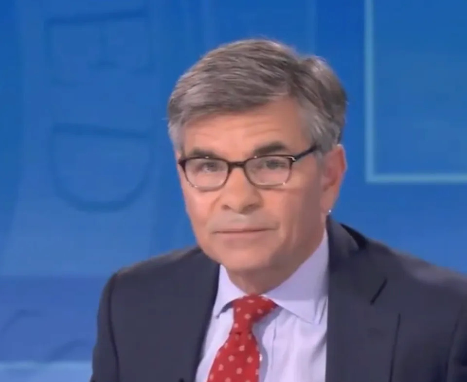 A news anchor in glasses and a suit with a red polka dot tie appears on a studio set