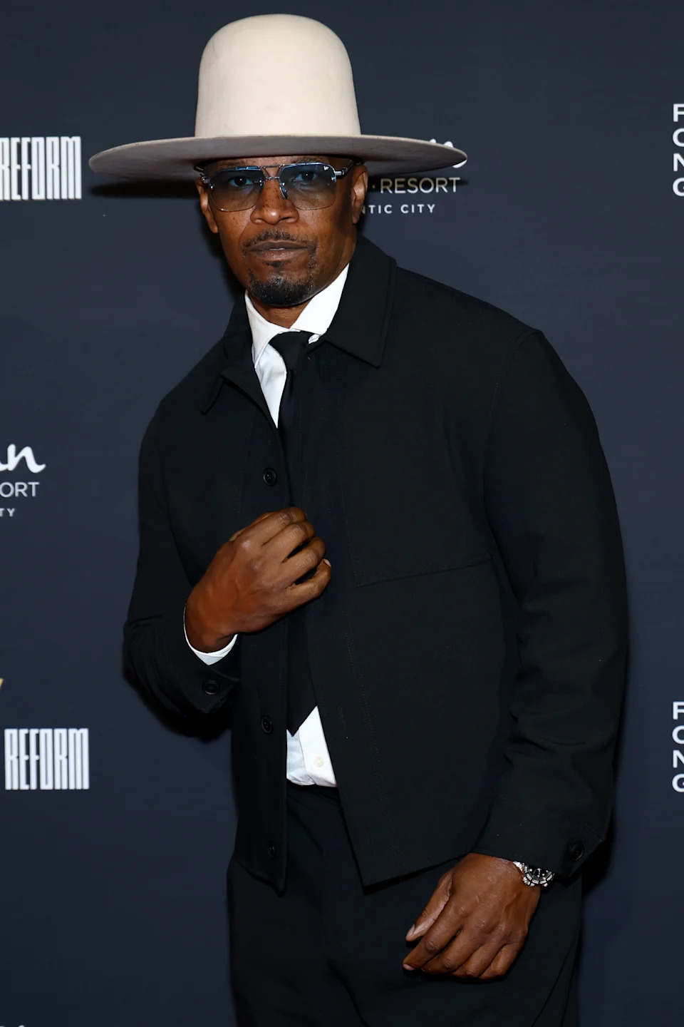 A man in a suit and wide-brimmed hat poses confidently at a formal event, wearing sunglasses and a stylish tie