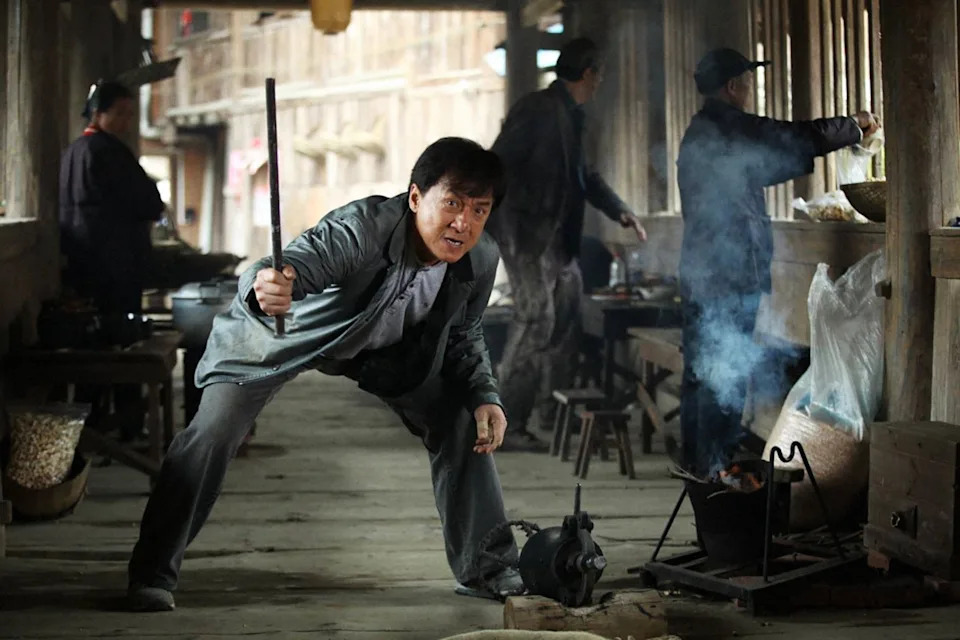 Person holding a stick, in a dramatic pose, surrounded by others cooking in a rustic kitchen setting