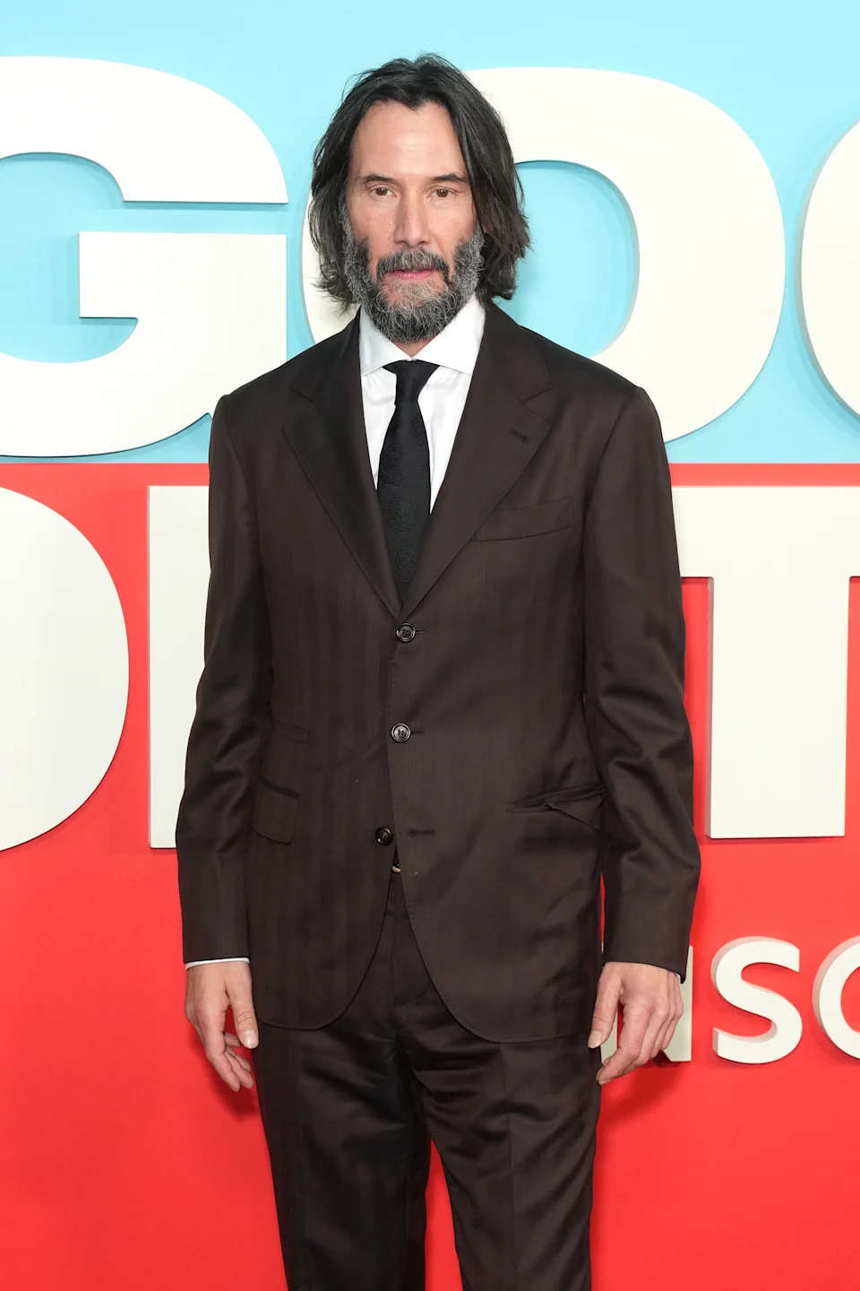 A man in a dark suit with a white shirt and black tie stands on a red carpet with large text in the background