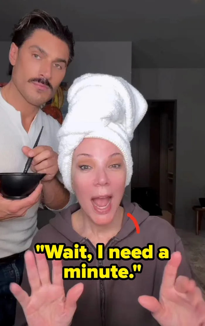 A person with a towel on their head smiling and posing with a man holding a bowl and brush, suggesting a beauty or skincare routine