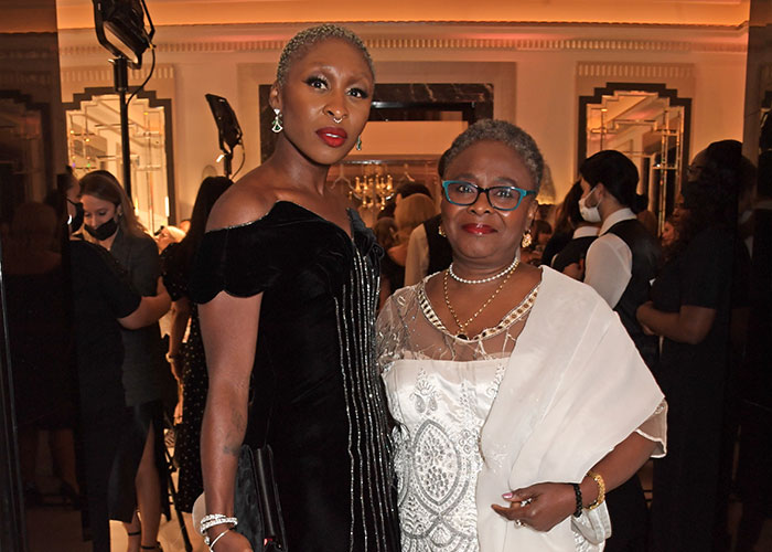 Cynthia Erivo in a black dress and another woman in white, attending an event with guests and photographers in the background.