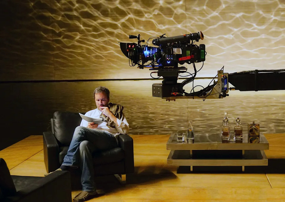 Person seated on a sofa, reading a script. A film camera is set up nearby on a robotic arm, suggesting a movie or TV production setting