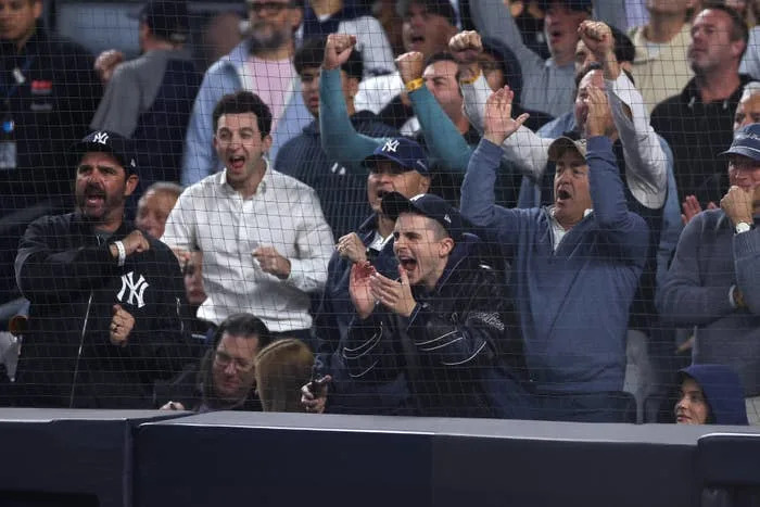 Excited crowd at a baseball game cheering behind a protective net, featuring fans in Yankees apparel