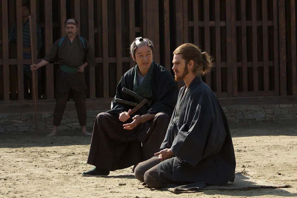 Three individuals in traditional attire, two sitting and one standing, in a historic setting. They appear to be engaged in a thoughtful conversation