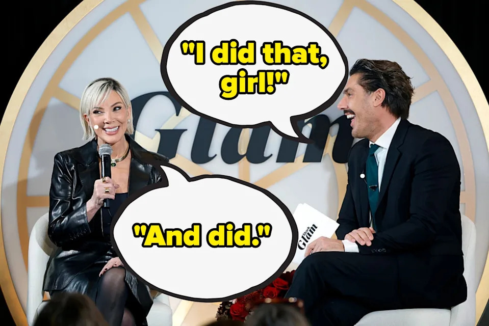 Two people sitting and smiling on stage during a talk show. The woman is in a leather jacket and holding a mic. The man is in a suit, holding papers