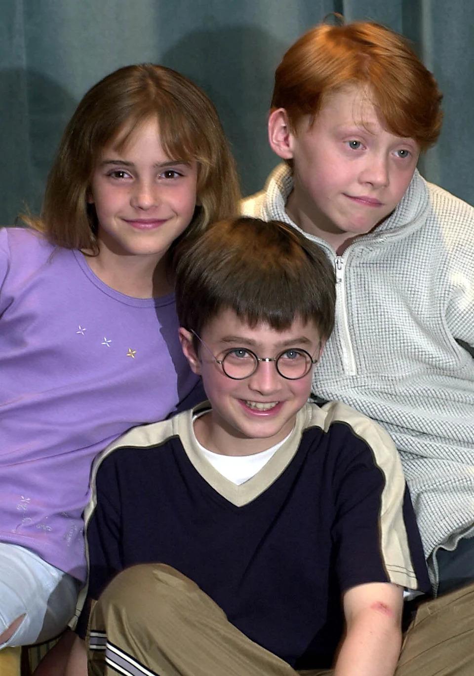 Three young actors from a famous film series smile for a photo. One wears a casual jacket, another a purple top, and the third has round glasses