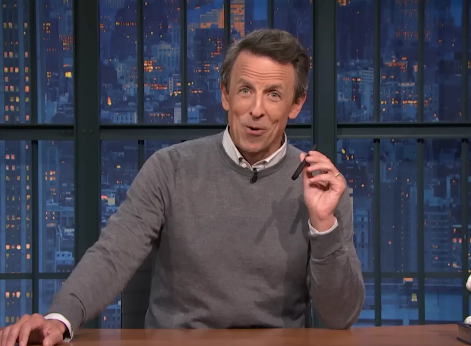 Person in a studio setting speaks animatedly, holding a pen, wearing a casual sweater over a collared shirt