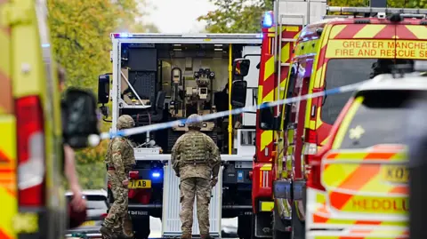 PA Media Members of the army in military fatigues stand outside a van with a bomb disposal robot seen inside. Police vehicles and police tape are seen in the foreground. 