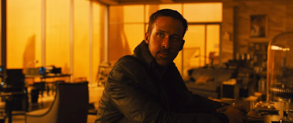A man in a futuristic setting sits at a table, wearing a dark jacket. The room has a warm, dim light with modern furniture visible in the background