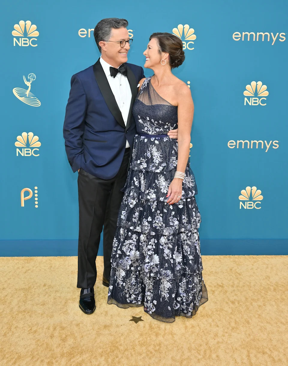 Stephen Colbert in a tuxedo and Evelyn McGee Colbert wears an elegant, floral-patterned, one-shoulder gown on the red carpet