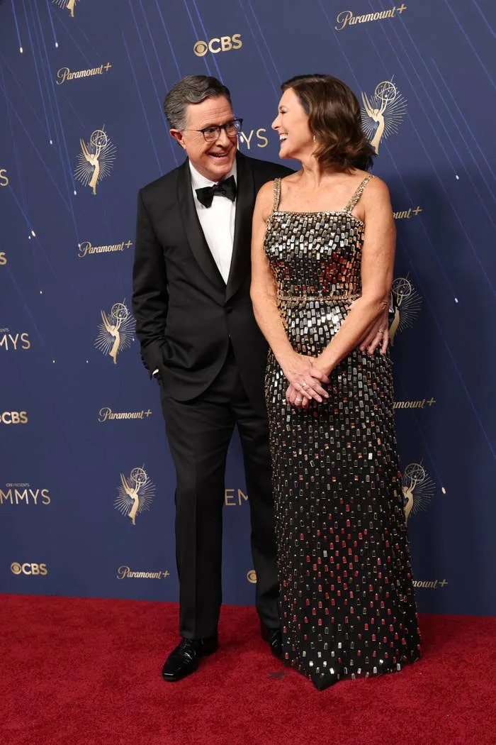 Stephen Colbert  in a tuxedo and Evelyn McGee in a sequin dress share a smile on the red carpet at a celebrity event
