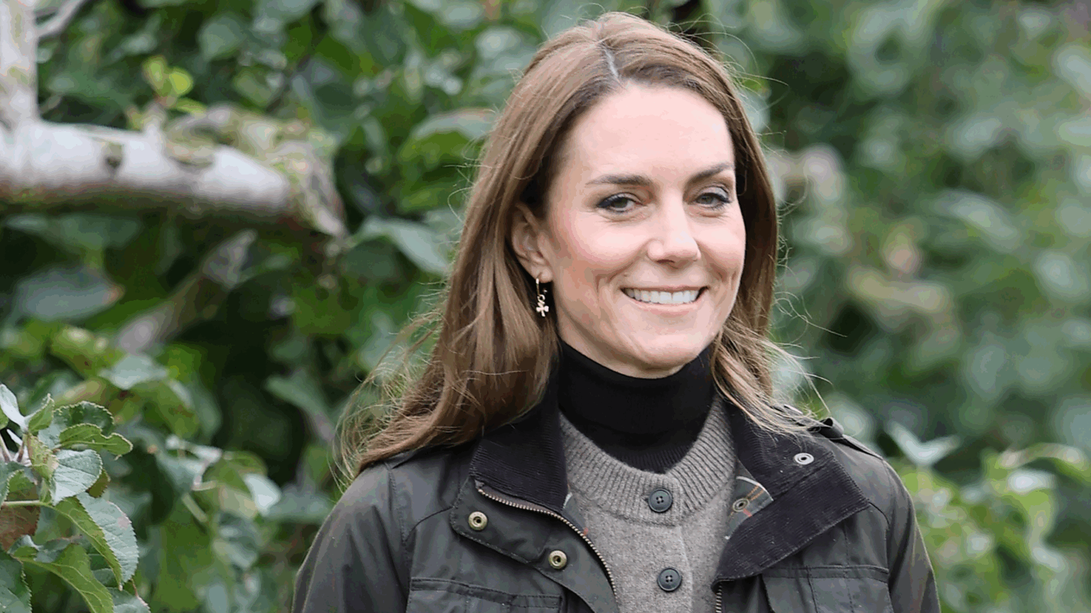 Kate Middleton wearing a green jacket and brown cardigan in front of trees