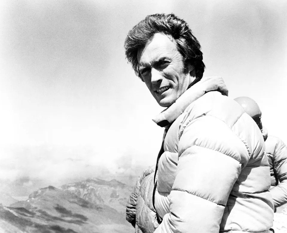 A person in a puffy jacket stands against a mountain backdrop, looking at the camera